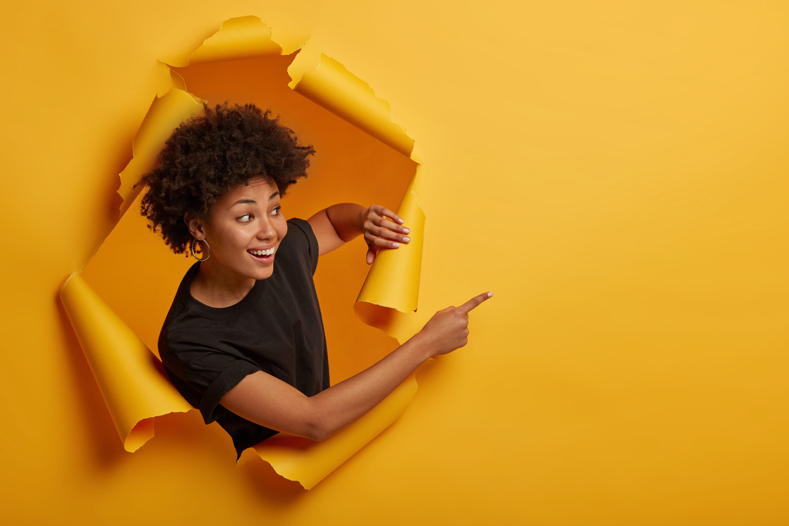Joyous friendly looking smiling girl points aside with happy expression, toothy smile, pleased to show awesome advertisement, dressed casually says use copy space wisely gestures through ripped paper